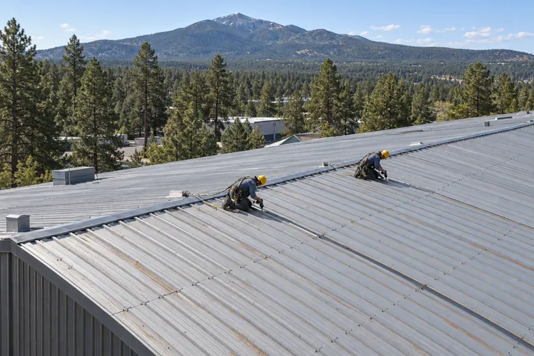 Two roofers replacing fasteners along the panel runs of an exposed-fastener metal warehouse roof